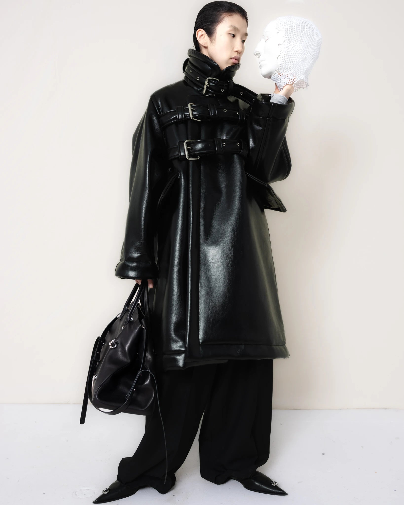 Person-wearing-a-black-shearling-oversized-coat-with- strap-and-buckle-details-worn-over-oversized-black-trousers-paired-with-a-large-black-leather-bag-and-black-pointed-leather-shoes-looking-at-a-white-mask-held-in-their-other-hand-full-body-front-view