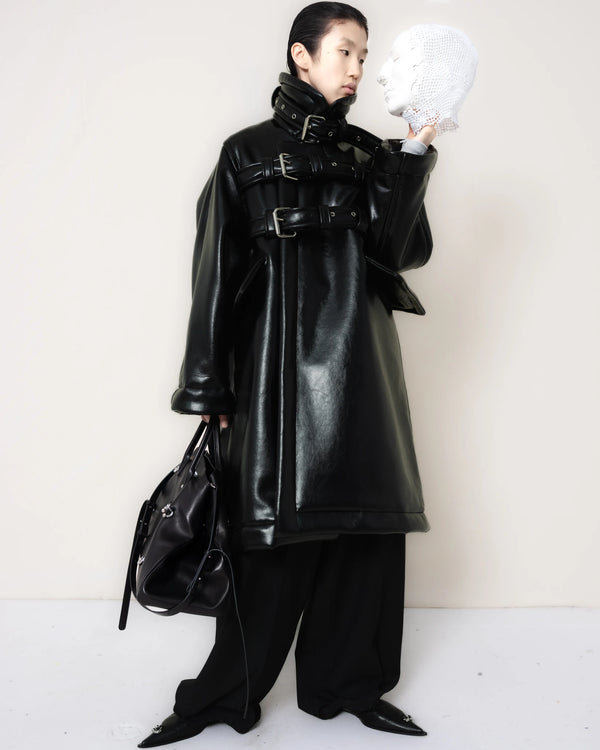 Person-wearing-a-black-shearling-oversized-coat-with- strap-and-buckle-details-worn-over-oversized-black-trousers-paired-with-a-large-black-leather-bag-and-black-pointed-leather-shoes-looking-at-a-white-mask-held-in-their-other-hand-full-body-front-view