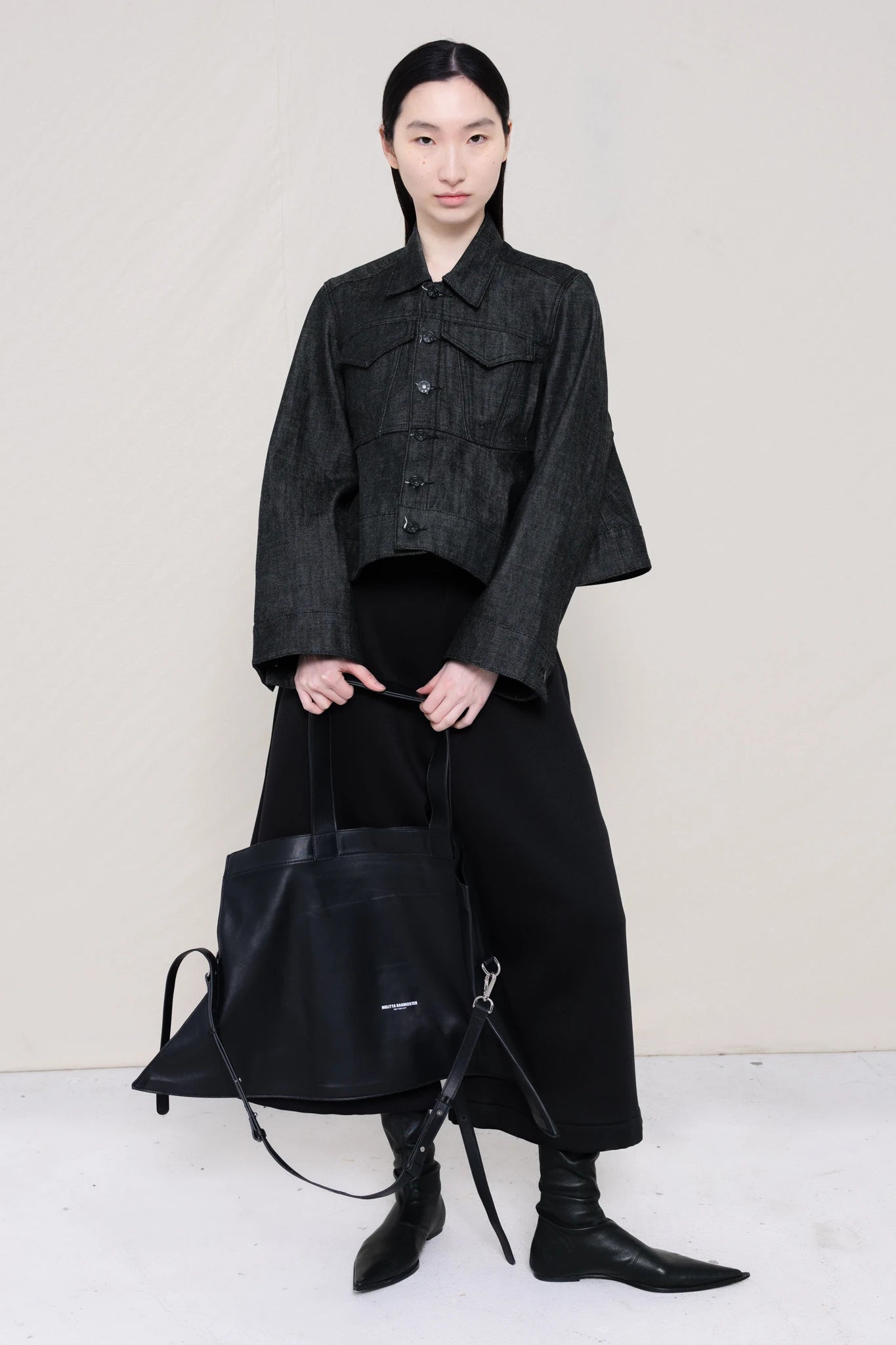 PERSON-WEARING-WASHED-CROPPED-BLACK-DENIM-JACKET-WITH-BLACK-PANTS-AND-FLAT-POINTY-BOOTS-HOLDING-A-LARGE-POINTED-PURSE-FRONT-VIEW