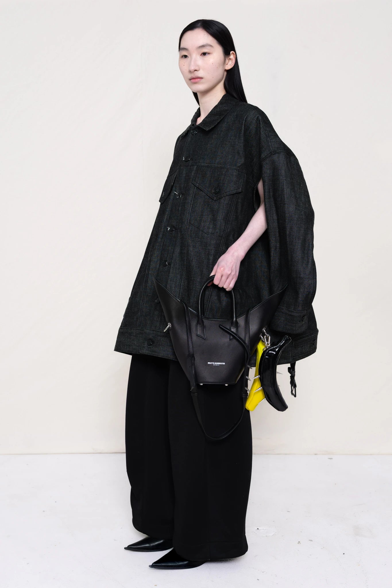 PERSON-WEARING-WASHED-BLACK-DENIM-JACKET-WITH-UNDERARM-HOLES-FOR-VERSITILE-WEARING-WITH-BLACK-TAILORED-PANTS-AND-FLAT-POINTY-BOOTS-HOLDING-A-POINTED-PURSE-WITH-TWO-BANANA-TOTES-CLIPPED-ON-THREE-QUARTER-VIEW