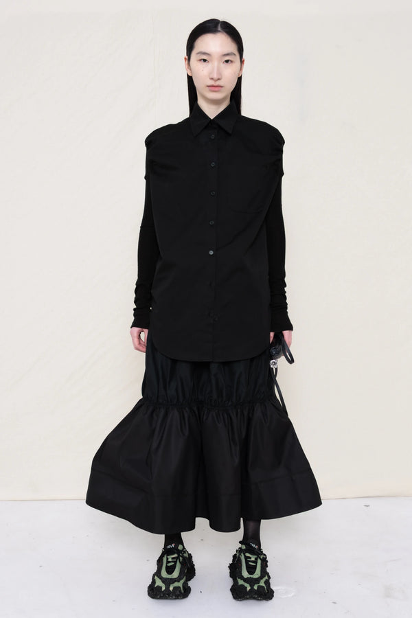 woman-holding-a-black-leather-banana-bag-with-stap-wearing-a-slim-black-collared-button-down-shirt-with-ruched-shoulder-detail-over-a-ruched-black-nylon-skirt-and-custom-shoes-against-a-plain-background-full-body-front-view