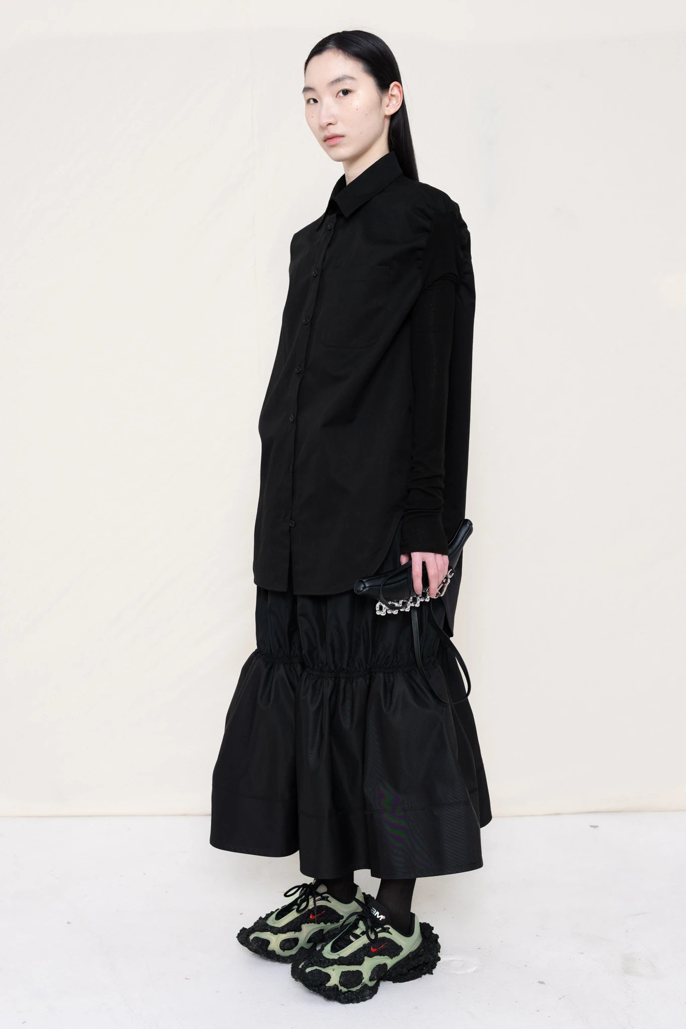 woman-holding-a-black-leather-banana-bag-with-stap-wearing-a-slim-black-collared-button-down-shirt-with-ruched-shoulder-detail-over-a-ruched-black-nylon-skirt-and-custom-shoes-against-a-plain-background-full-body-three-quarter-view