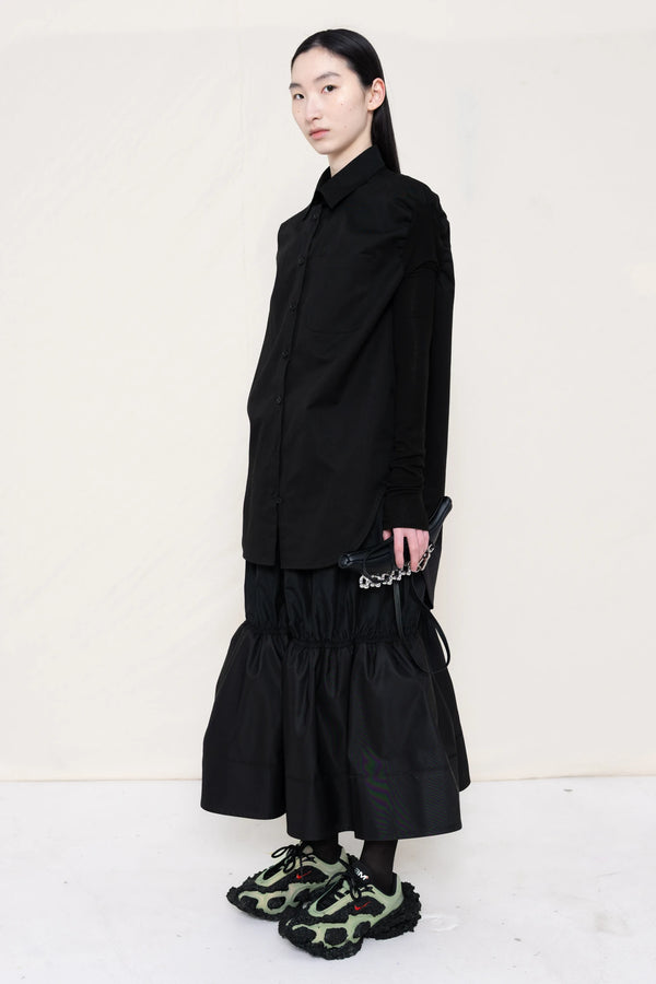 woman-holding-a-black-leather-banana-bag-with-stap-wearing-a-slim-black-collared-button-down-shirt-with-ruched-shoulder-detail-over-a-ruched-black-nylon-skirt-and-custom-shoes-against-a-plain-background-full-body-three-quarter-view