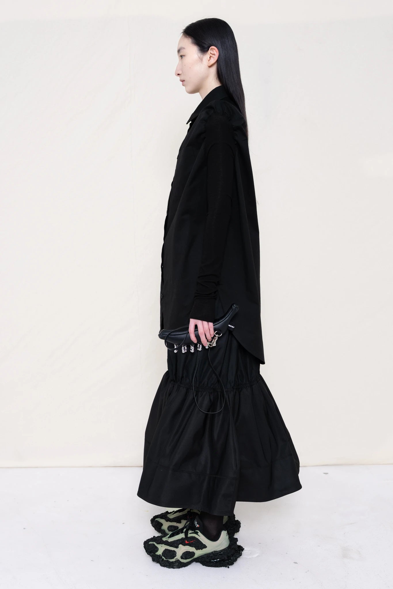 PERSON-WEARING-BLACK-SLIM-SLEEVE-BUTTON-DOWN-WITH-BLACK-RUCHED-SKIRT-AND-CUSTOMIZED-SNEAKERS-HOLDING-BLACK-BANANA-TOTE-SIDE-VIEW