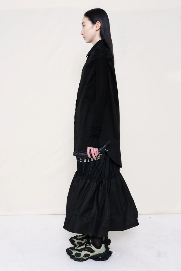 PERSON-WEARING-BLACK-SLIM-SLEEVE-BUTTON-DOWN-WITH-BLACK-RUCHED-SKIRT-AND-CUSTOMIZED-SNEAKERS-HOLDING-BLACK-BANANA-TOTE-SIDE-VIEW