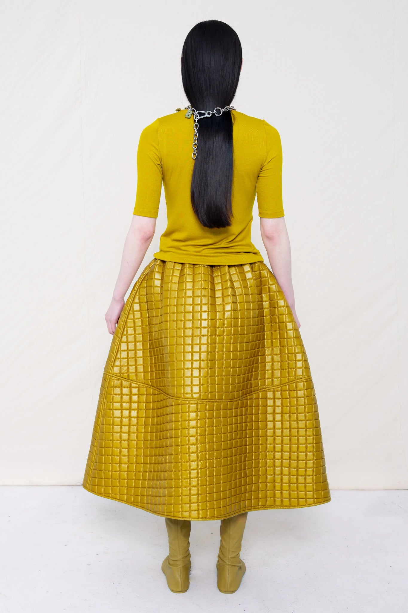 PERSON-WEARING-GOLD-YELLOW-SHORT-SLEEVE-MOCK-NECK-TOP-WITH-MATCHING-QUILTED-A-LINE-SKIRT-ACCESSORIZED-WITH-MB-CHARM-NECKLACE-AND-MUD-COLORED-POINTY-FLAT-BOOTS-BACK-VIEW