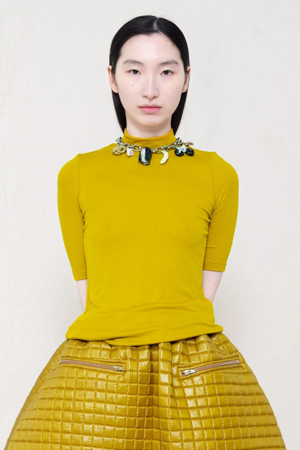 woman-wearing-A-shORT-SLEEVE-gold-BABY-MOCK-NECK-TOP-with-charm-chocker-over-a-quilted- foamy-gold-dropped-waisted-o-line-skirt-with-zipper pockets-against-a-plain-background-front-view