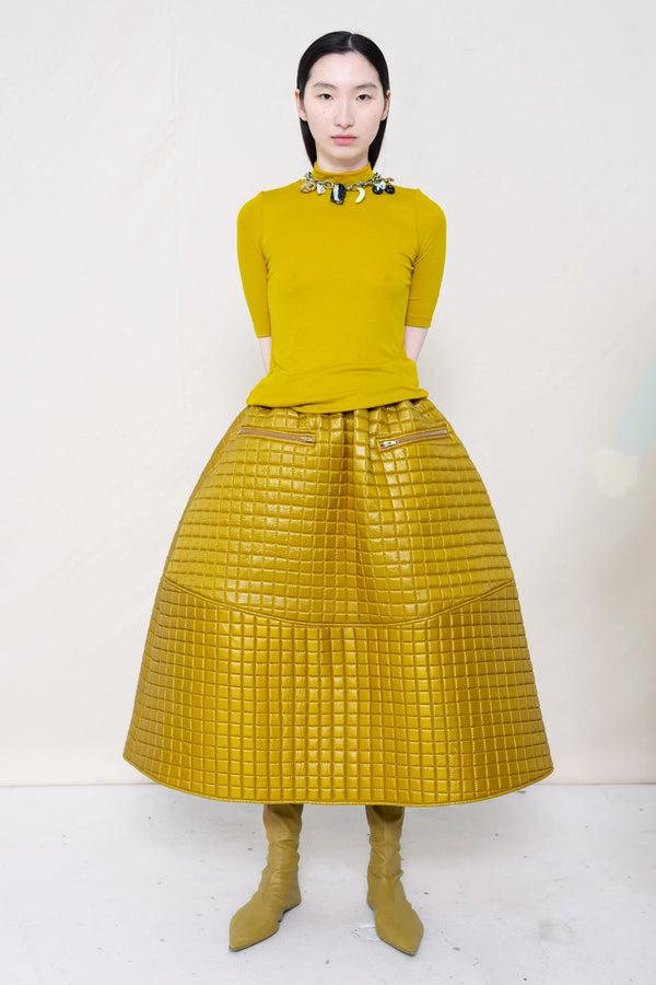 woman-wearing-a-short-sleeve-gold-baby-mock-neck-top-with-charm-choker-over-a-quilted-foamy-gold-dropped-waist-o-line-skirt-with-zipper-pockets-and-mustard-yellow-pointed-leather-boots-against-a-plain-background-full-body-front-view