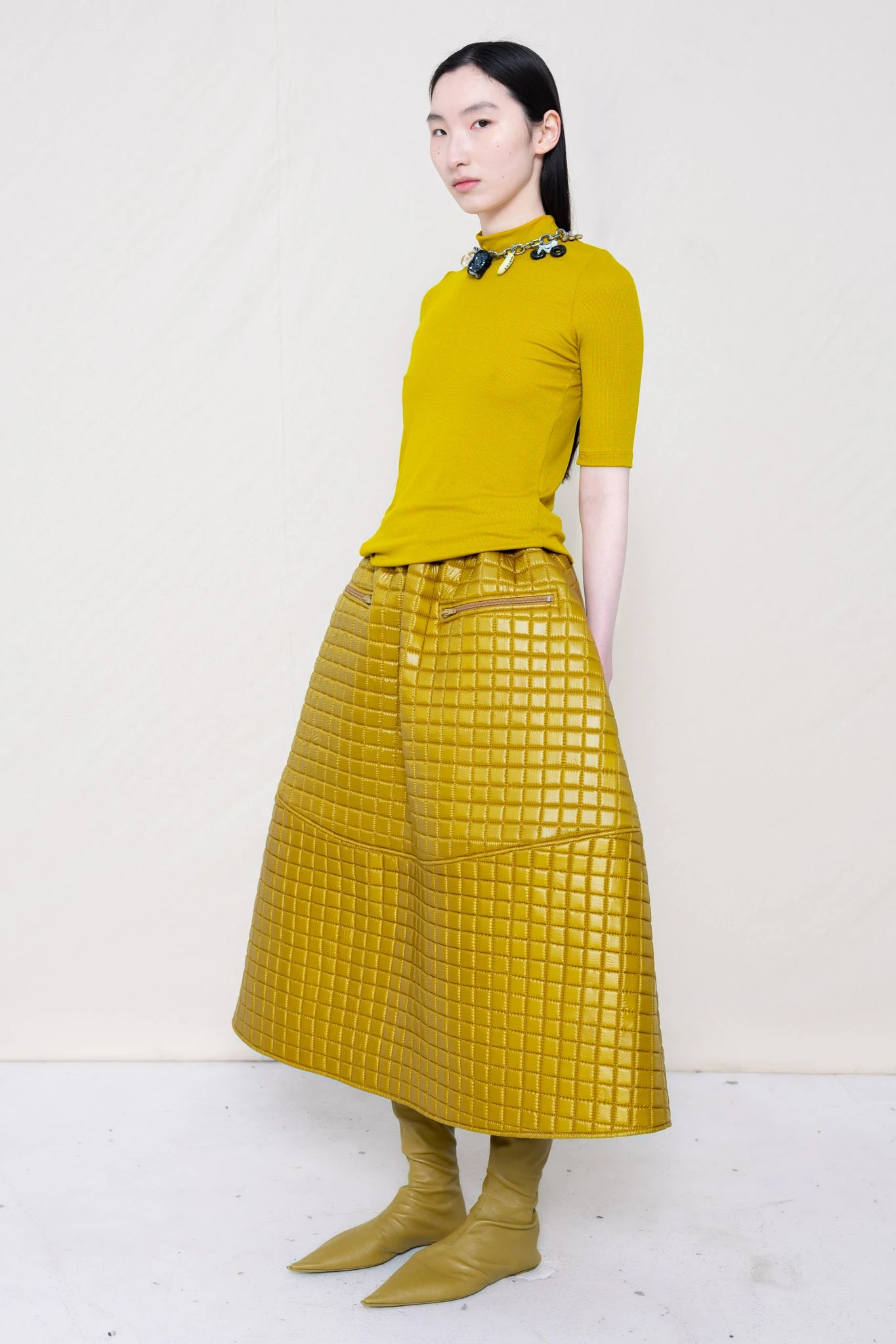 woman-wearing-a-short-sleeve-gold-baby-mock-neck-top-with-charm-choker-over-a-quilted-foamy-gold-dropped-waist-o-line-skirt-with-zipper-pockets-and-mustard-yellow-pointed-leather-boots-against-a-plain-background-full-body-three-quarter-view