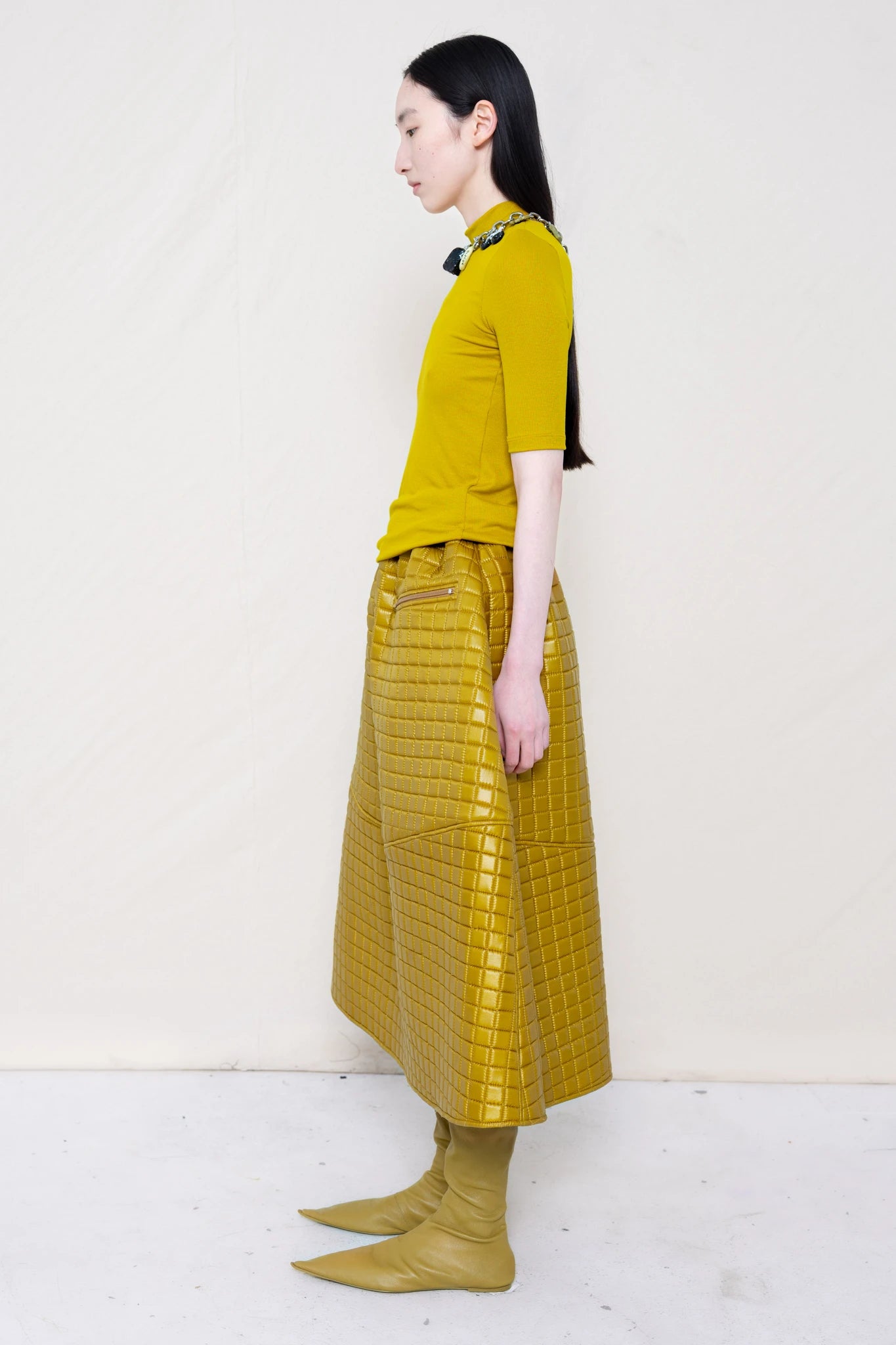 PERSON-WEARING-GOLD-YELLOW-SHORT-SLEEVE-MOCK-NECK-TOP-WITH-MATCHING-QUILTED-A-LINE-SKIRT-ACCESSORIZED-WITH-MB-CHARM-NECKLACE-AND-MUD-COLORED-POINTY-FLAT-BOOTS-SIDE-VIEW