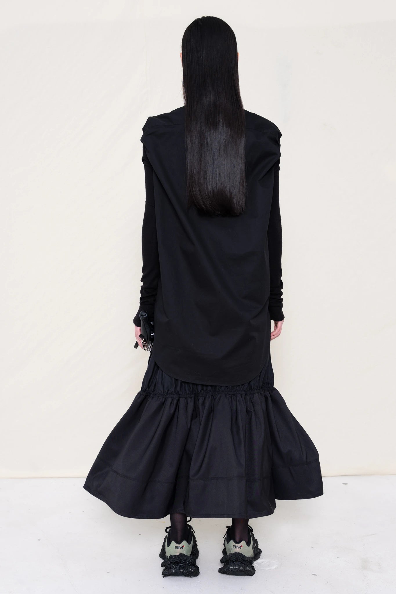 woman-holding-a-black-leather-banana-bag-with-stap-wearing-a-slim-black-collared-button-down-shirt-with-ruched-shoulder-detail-over-a-ruched-black-nylon-skirt-and-custom-shoes-against-a-plain-background-full-body-back-view