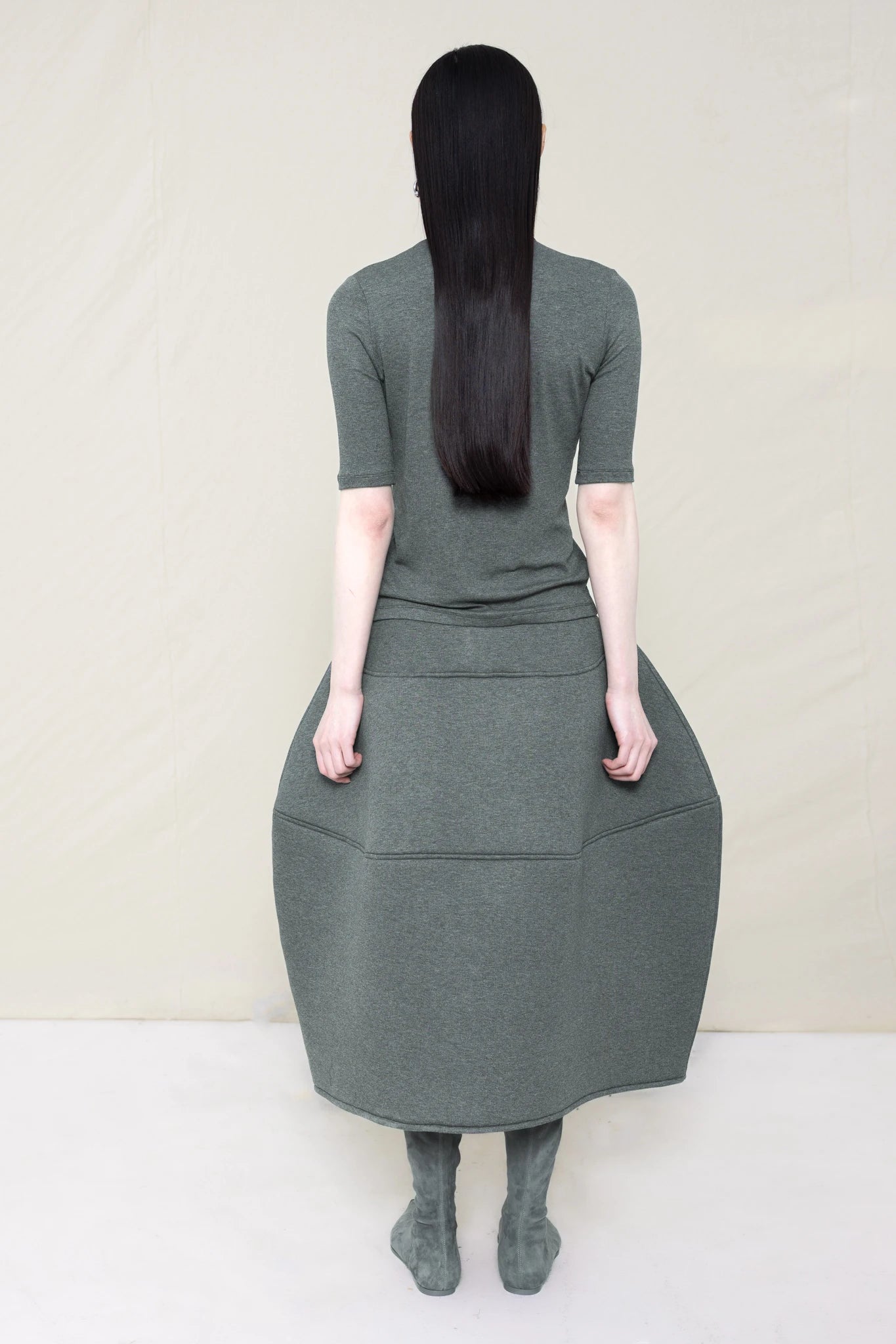 woman-wearing-A-shORT-SLEEVE-grey-BABY-MOCK-NECK-TOP-over-a-foamy-grey-dropped-waisted-o-line-skirt-with-grey-pointed-boots-against-a-plain-background-full-body-back-view