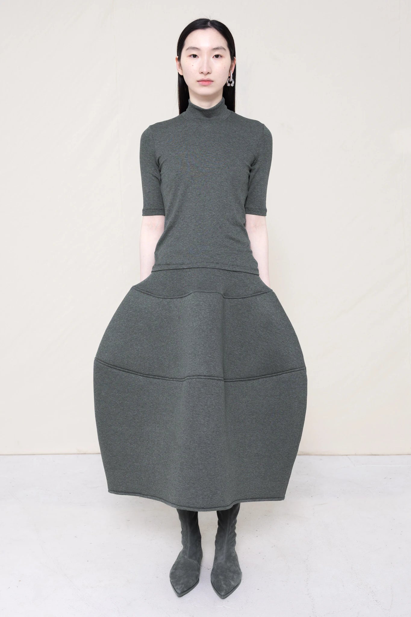 woman-wearing-A-shORT-SLEEVE-grey-BABY-MOCK-NECK-TOP-over-a-foamy-grey-dropped-waisted-o-line-skirt-with-grey-pointed-boots-against-a-plain-background-full-body-front-view