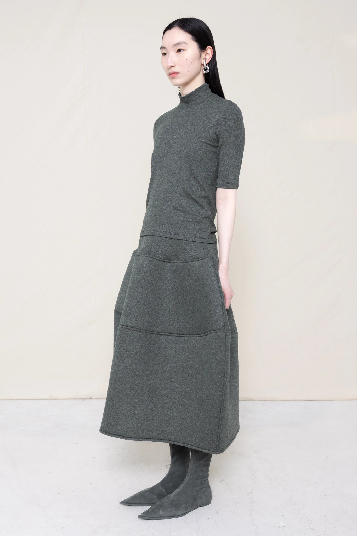 woman-wearing-A-shORT-SLEEVE-grey-BABY-MOCK-NECK-TOP-over-a-foamy-grey-dropped-waisted-o-line-skirt-with-grey-pointed-boots-against-a-plain-background-full-body-three-quarter-view
