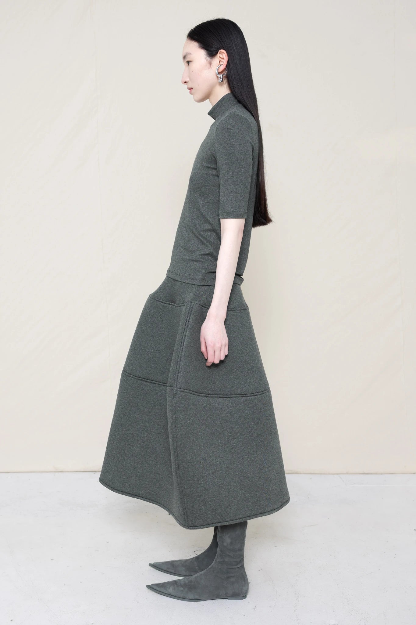 woman-wearing-A-shORT-SLEEVE-grey-BABY-MOCK-NECK-TOP-over-a-foamy-grey-dropped-waisted-o-line-skirt-with-grey-pointed-boots-against-a-plain-background-full-body-side-view