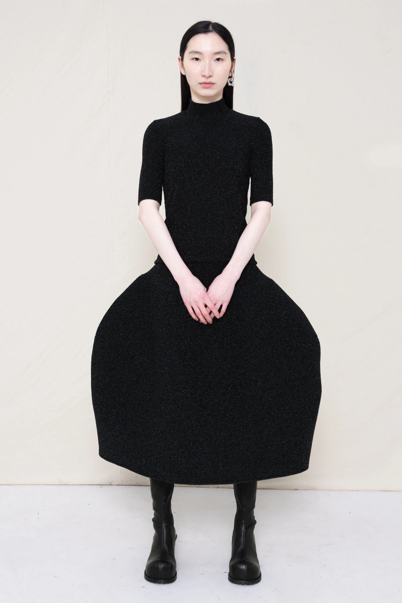 woman-wearing-a-short-sleeve-black-glitter-baby-mock-neck-top-over-a--foamy-black-glitter-dropped-waist-o-line-skirt-and-black-leather-boots-against-a-plain-background-full-body-front-view