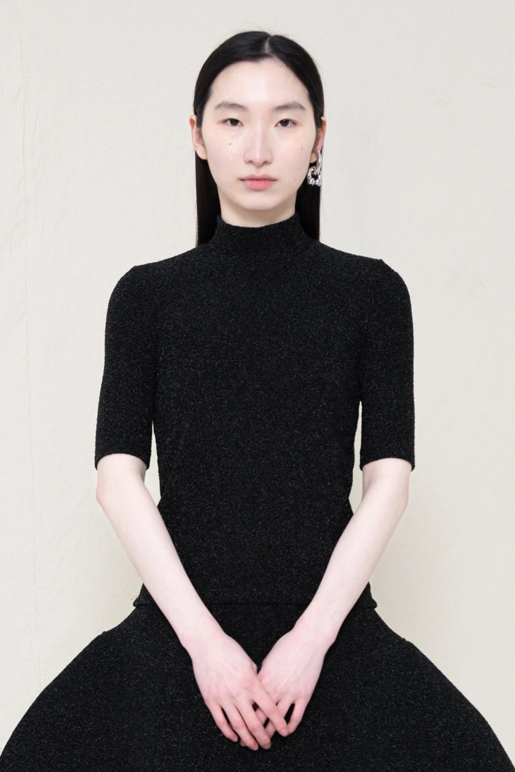 woman-wearing-a-short-sleeve-black-glitter-baby-mock-neck-top-over-a--foamy-black-glitter-dropped-waist-o-line-skirt-and-black-leather-boots-against-a-plain-background-front-view