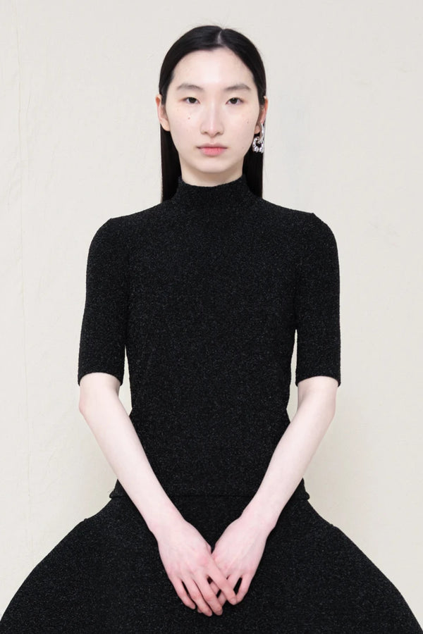 woman-wearing-a-short-sleeve-black-glitter-baby-mock-neck-top-over-a--foamy-black-glitter-dropped-waist-o-line-skirt-and-black-leather-boots-against-a-plain-background-front-view
