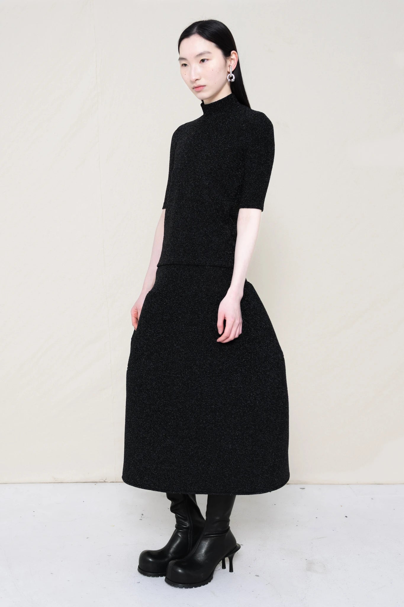 woman-wearing-a-short-sleeve-black-glitter-baby-mock-neck-top-over-a--foamy-black-glitter-dropped-waist-o-line-skirt-and-black-leather-boots-against-a-plain-background-full-body-three-quarter-view