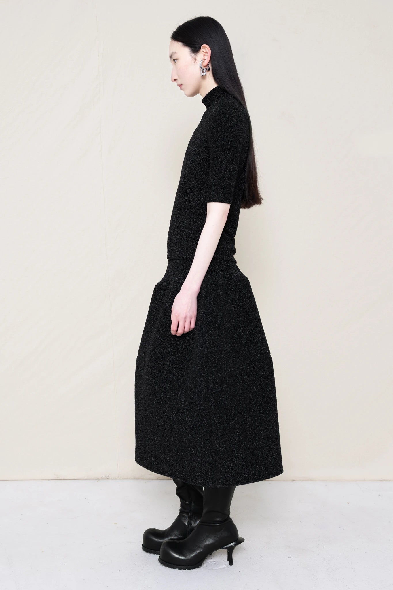 woman-wearing-a-short-sleeve-black-glitter-baby-mock-neck-top-over-a--foamy-black-glitter-dropped-waist-o-line-skirt-and-black-leather-boots-against-a-plain-background-full-body-side-view