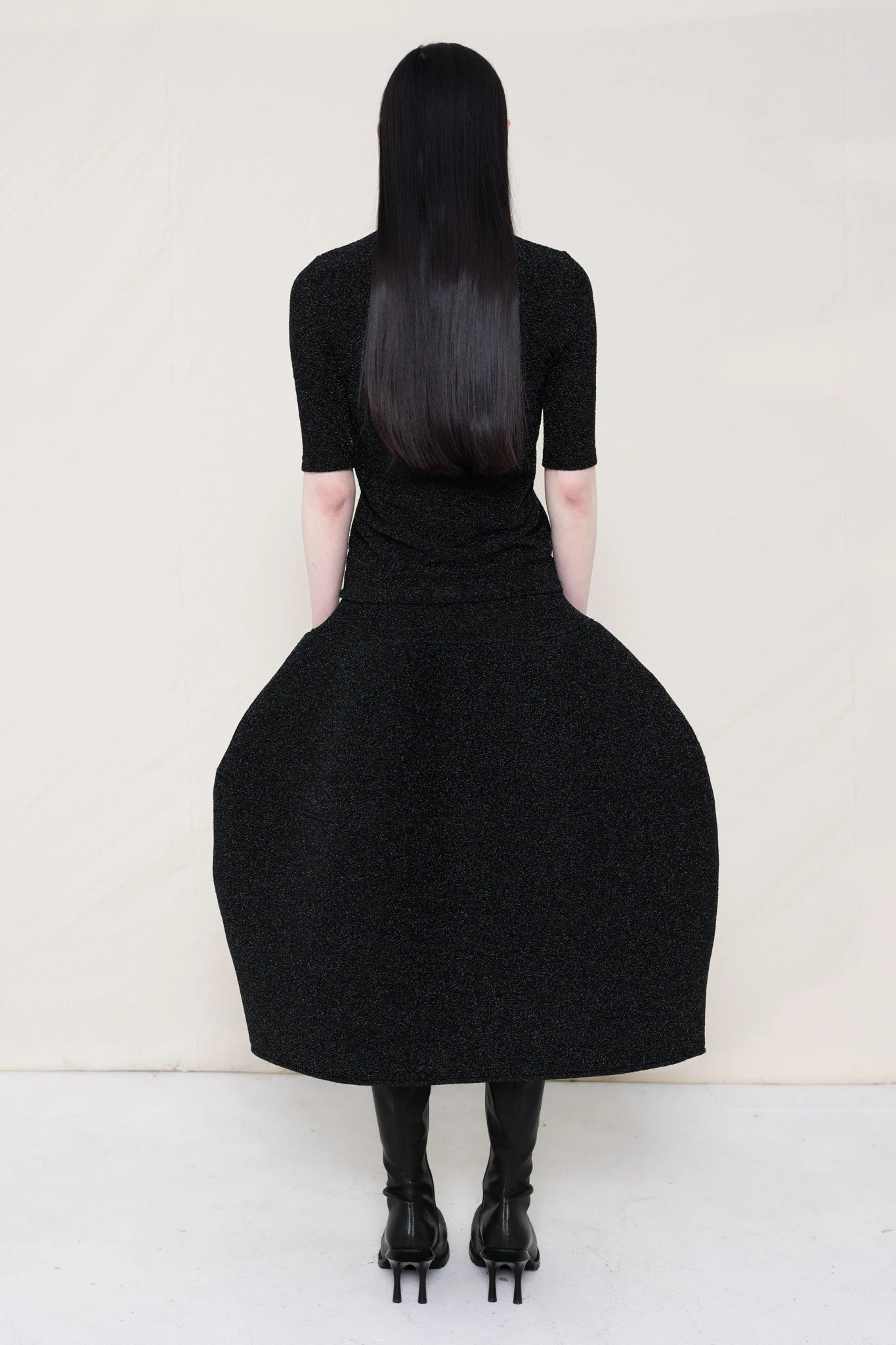 woman-wearing-a-short-sleeve-black-glitter-baby-mock-neck-top-over-a--foamy-black-glitter-dropped-waist-o-line-skirt-and-black-leather-boots-against-a-plain-background-full-body-back-view