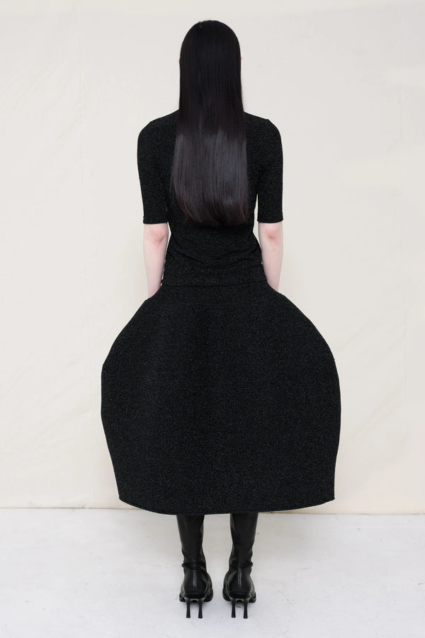 woman-wearing-a-short-sleeve-black-glitter-baby-mock-neck-top-over-a--foamy-black-glitter-dropped-waist-o-line-skirt-and-black-leather-boots-against-a-plain-background-full-body-back-view