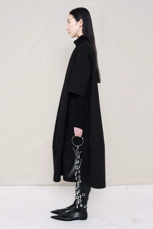 woman-holding-a-black-leather-studded-banana-wearing-a-calf-length-black-a-line-boatneck-dress-with-mid-length-sleeves-worn-over-a-black-turtle-neck-worn-with-black-pointed-studded-leather-boots-against-a-plain-background-full-body-side-view