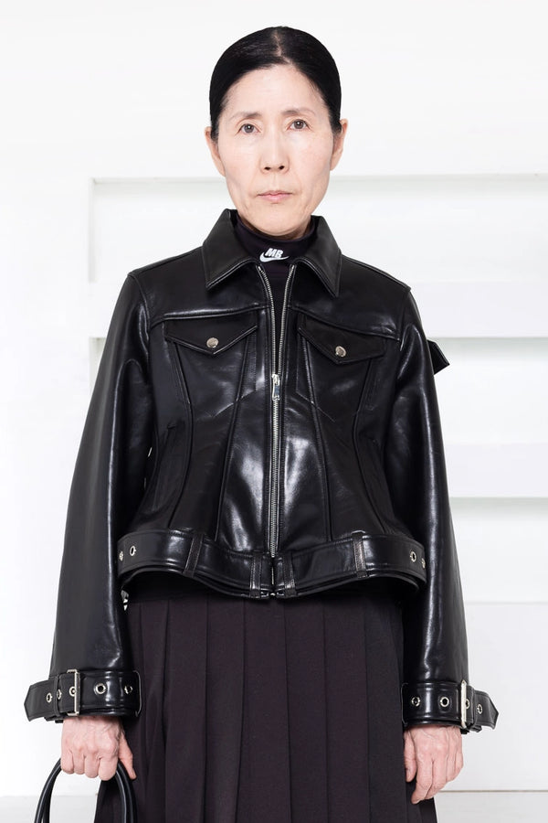 A woman wearing a cropped black leather jacket with silver-tone hardware and a high collar, paired with a dark skirt, standing against a white paneled background.