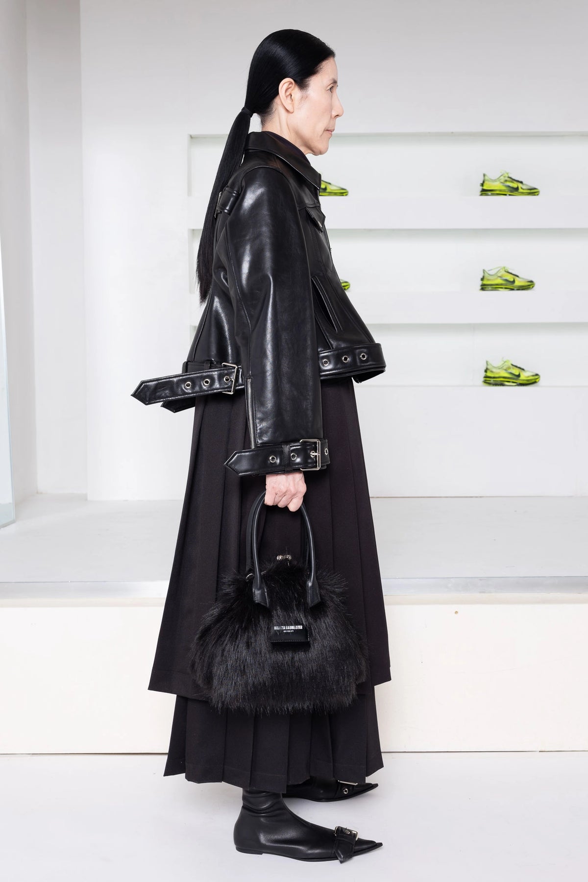 A model in a side profile wearing a black leather jacket with wide lapels and belt details, a long black pleated skirt, holding a black fur-trimmed handbag, in a minimalist white room with green shoes on display.