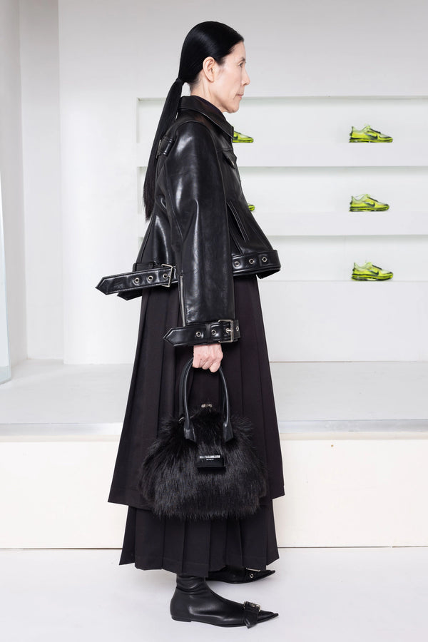 A model in a side profile wearing a black leather jacket with wide lapels and belt details, a long black pleated skirt, holding a black fur-trimmed handbag, in a minimalist white room with green shoes on display.