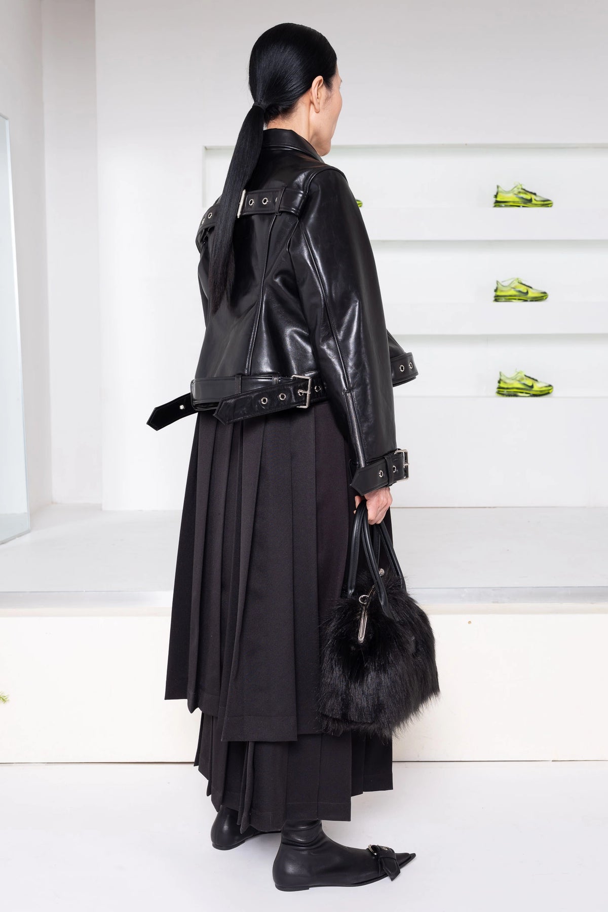 A model shown from the back wearing a black leather jacket with a wide strap and belt, a long black pleated skirt, black boots, and holding a black fur-trim handbag in a minimalist white room.