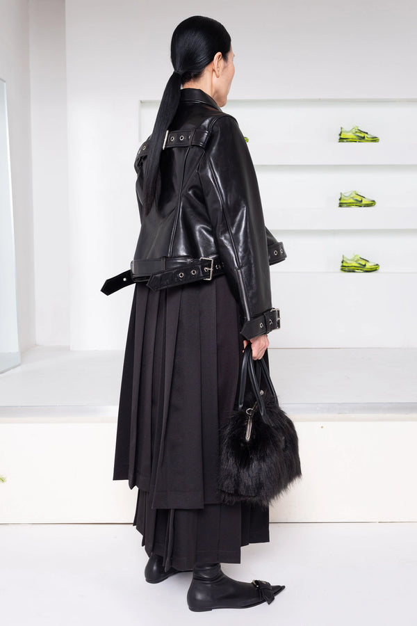 A model shown from the back wearing a black leather jacket with a wide strap and belt, a long black pleated skirt, black boots, and holding a black fur-trim handbag in a minimalist white room.