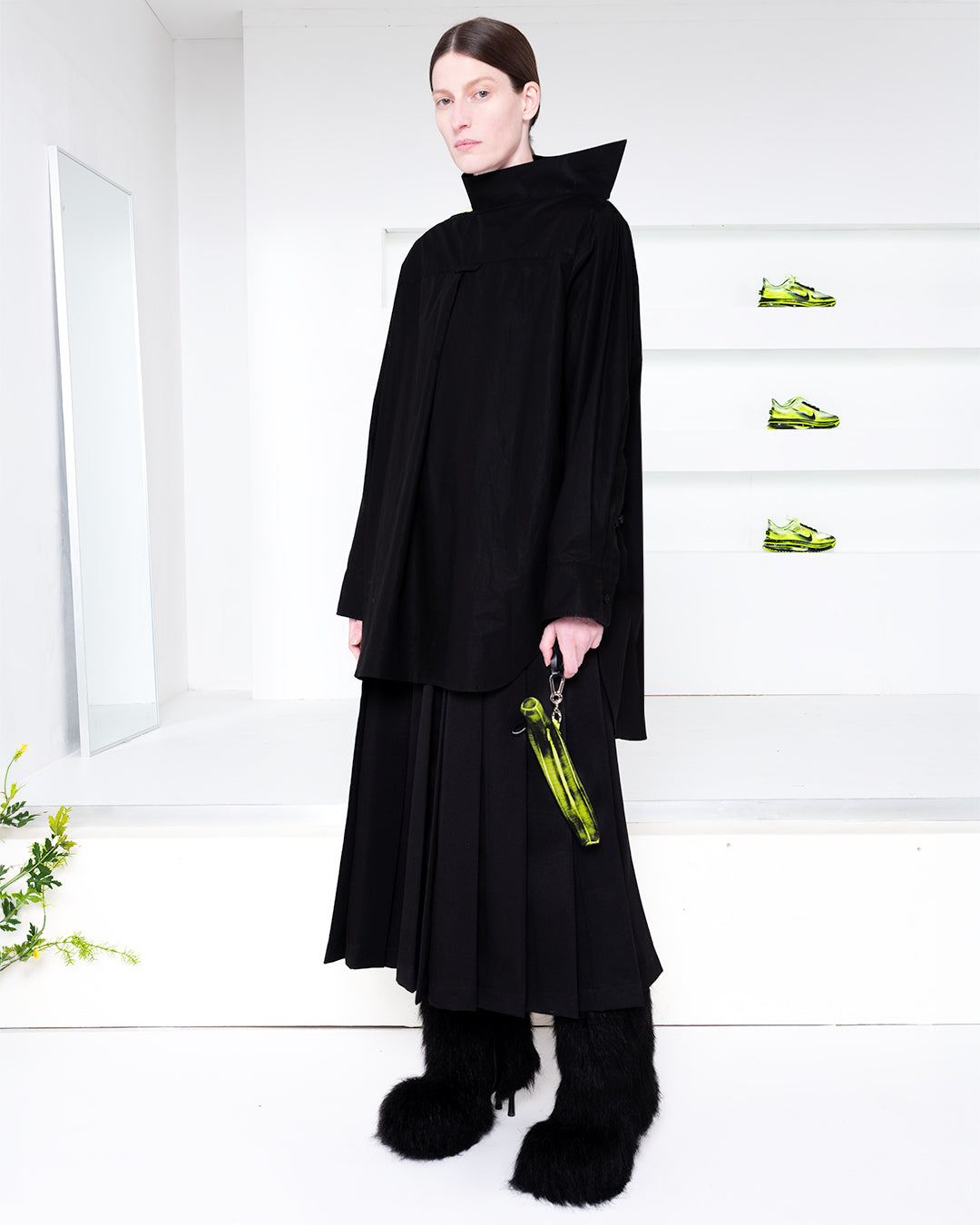 Model wearing all black outfit in a white room with green shoes in shelves in the back