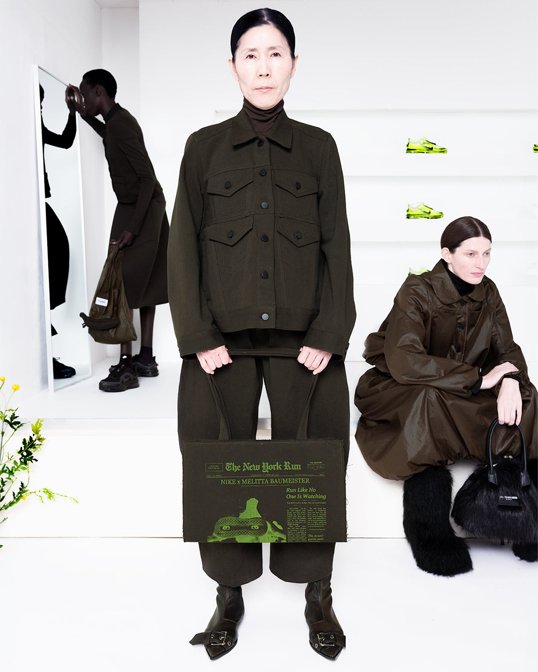 Three models in brown outfits in a white room with green shoes in shelves in the back