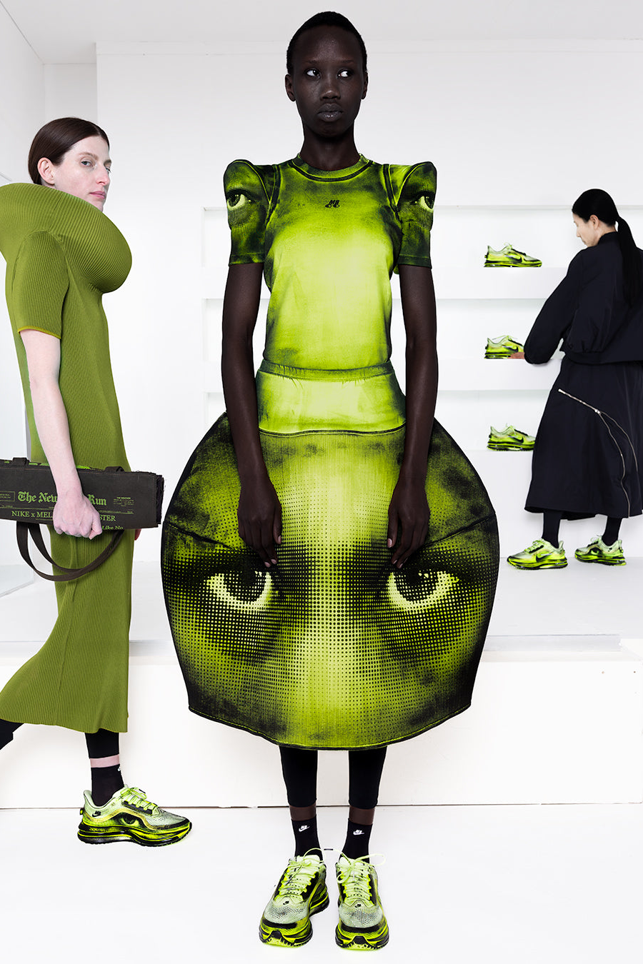Model wearing a green dress with eye design on a white staircase.