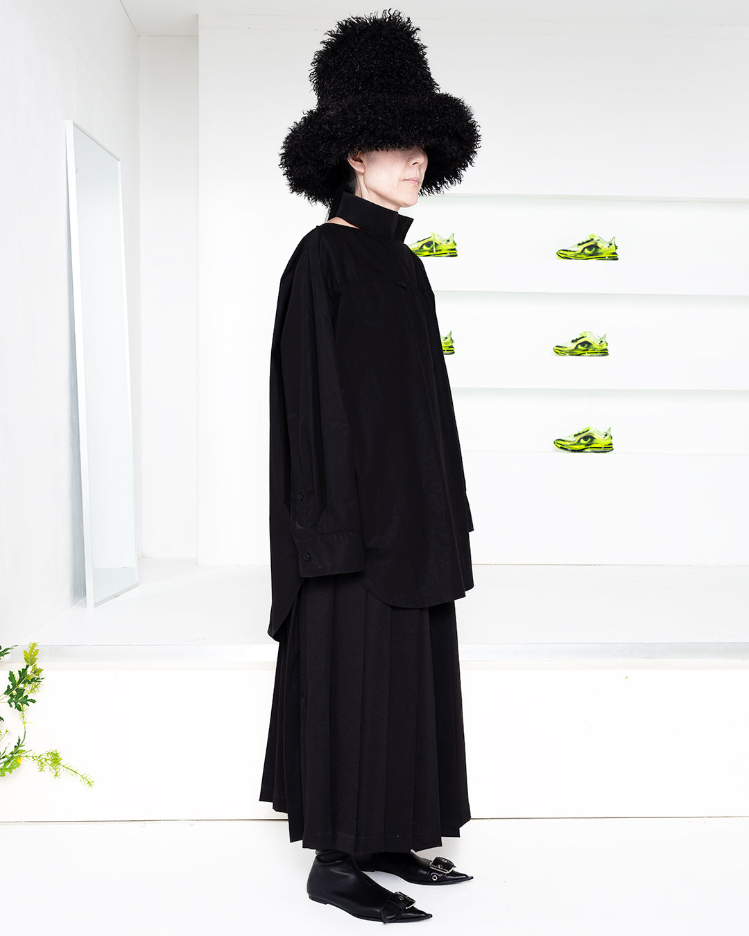 Model wearing a black outfit in a white room with green shoes in shelves in the back