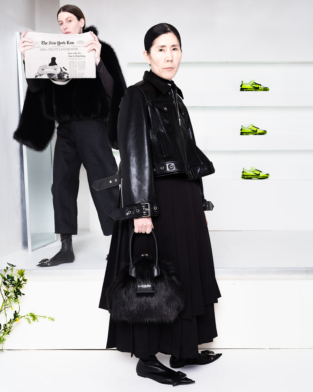 Two models  in a white room with green shoes in shelves in the back