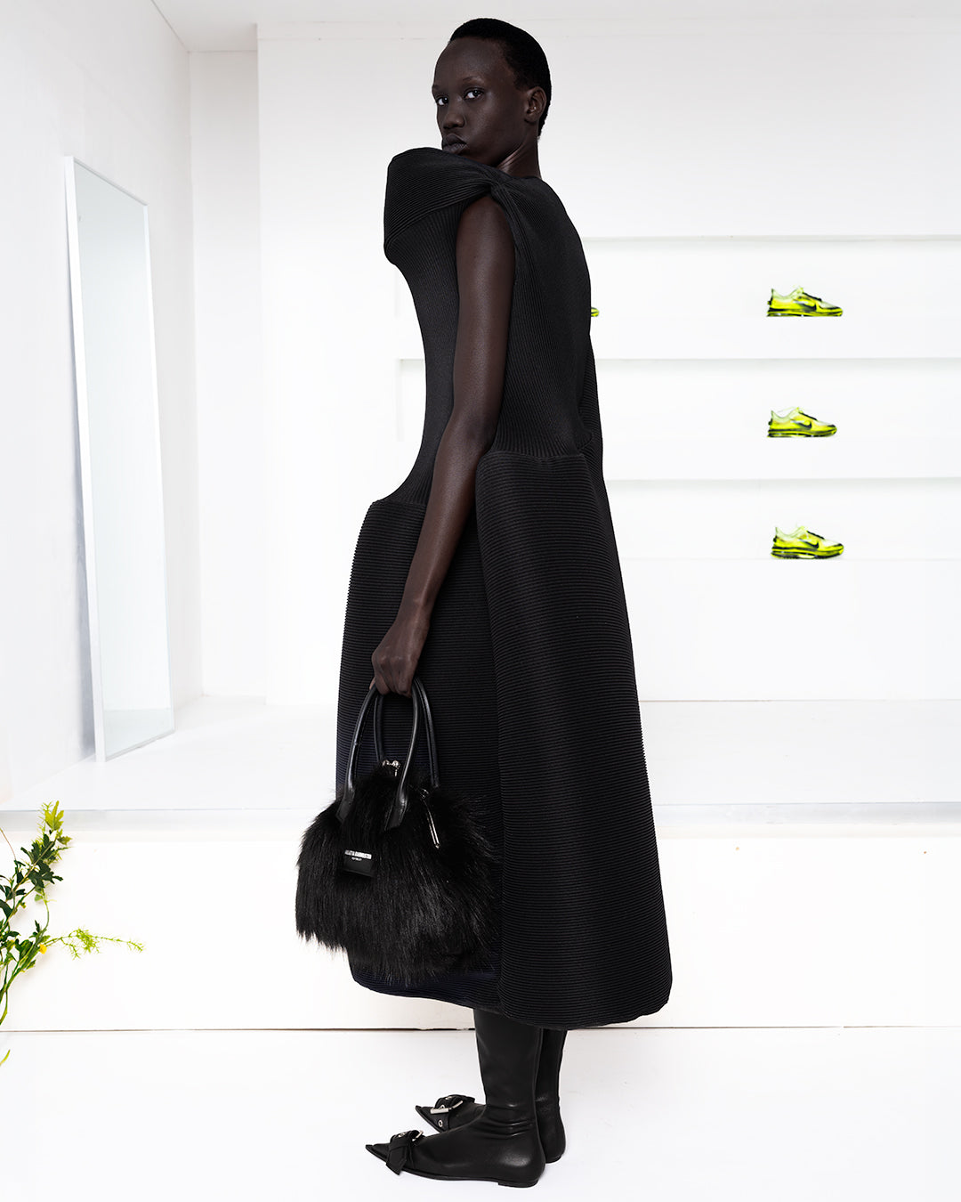 Model in a pleated dress in a white room with green shoes in shelves in the back