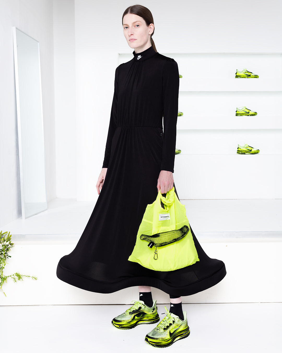Model wearing a black dress with yellow bag and shoes in a white room with green shoes in shelves in the back