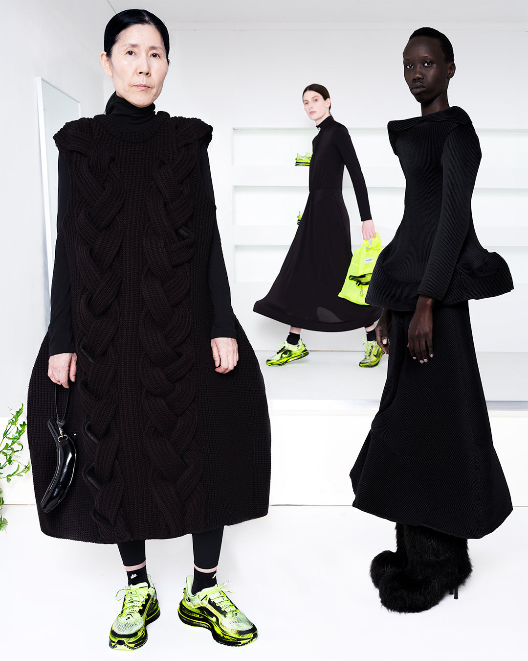 Three models wearing black in a white room with green shoes in shelves in the back
