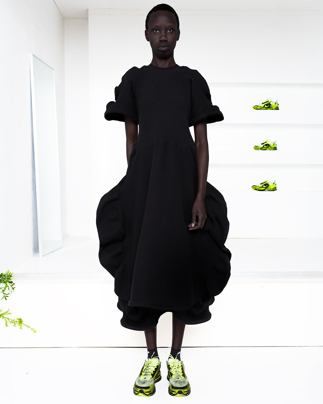 Model wearing a black dress in a white room with green shoes in shelves in the back