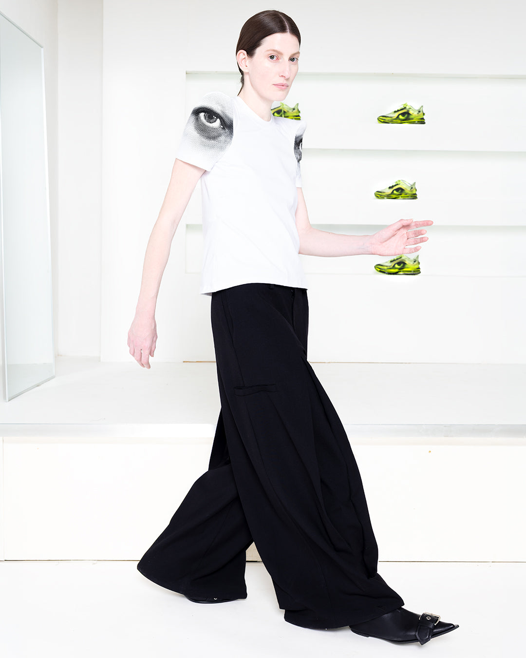 Model wearing white tee and black pants in a white room with green shoes in shelves in the back