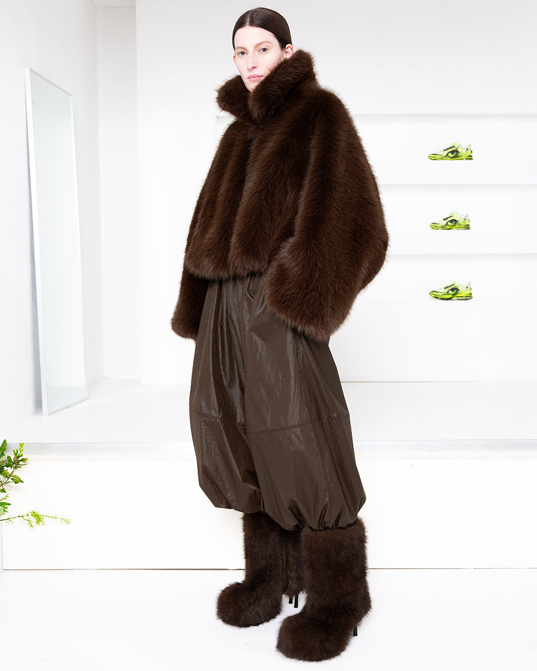 Model wearing brown fuzzy jacket in a white room with green shoes in shelves in the back