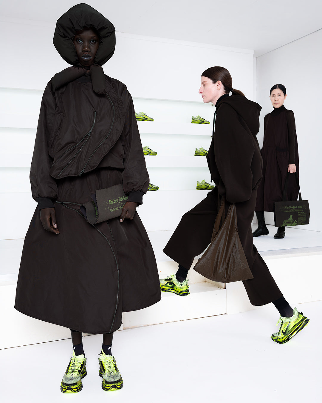 Three models in a white room with green shoes in shelves in the back