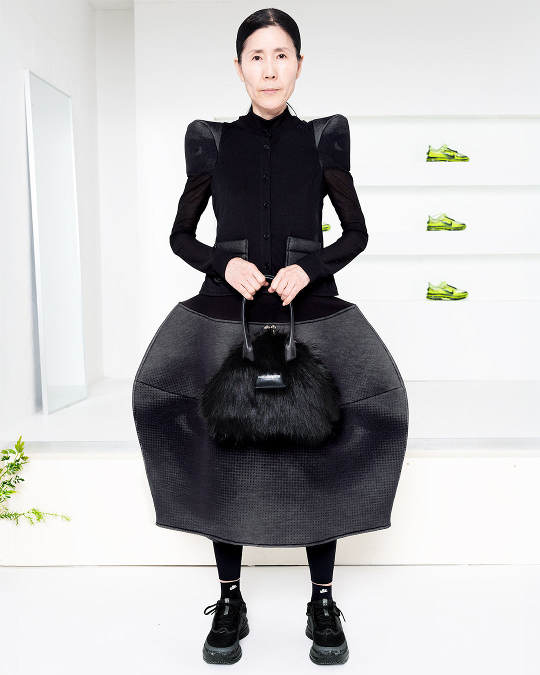 Model wearing a black dress in a white room with green shoes in shelves in the back