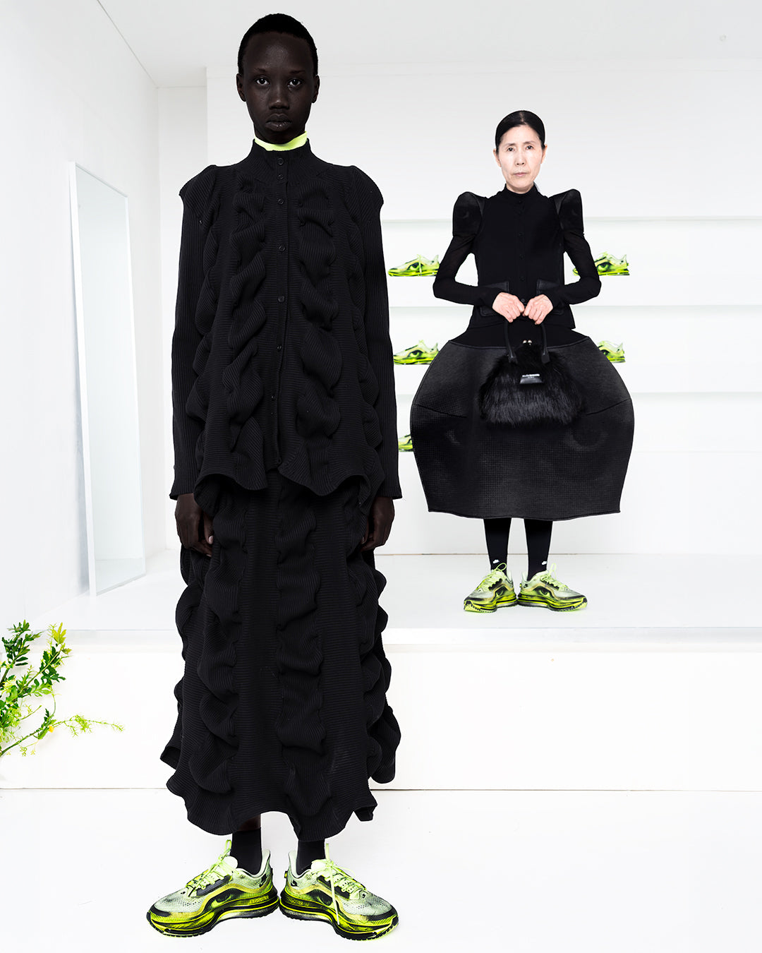 Two models wearing black in a white room with green shoes in shelves in the back
