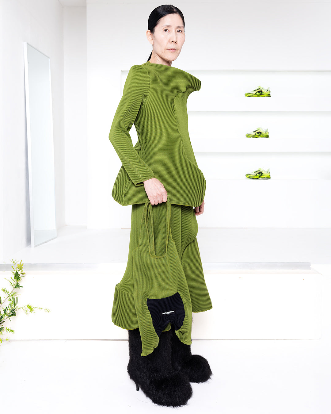 Model wearing pleated green outfit in a white room with green shoes in shelves in the back