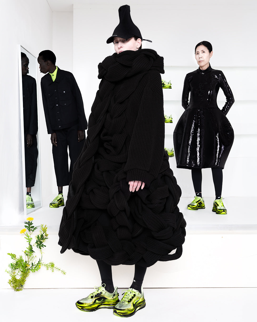 Three models in black outfits in a white room with green shoes in shelves in the back