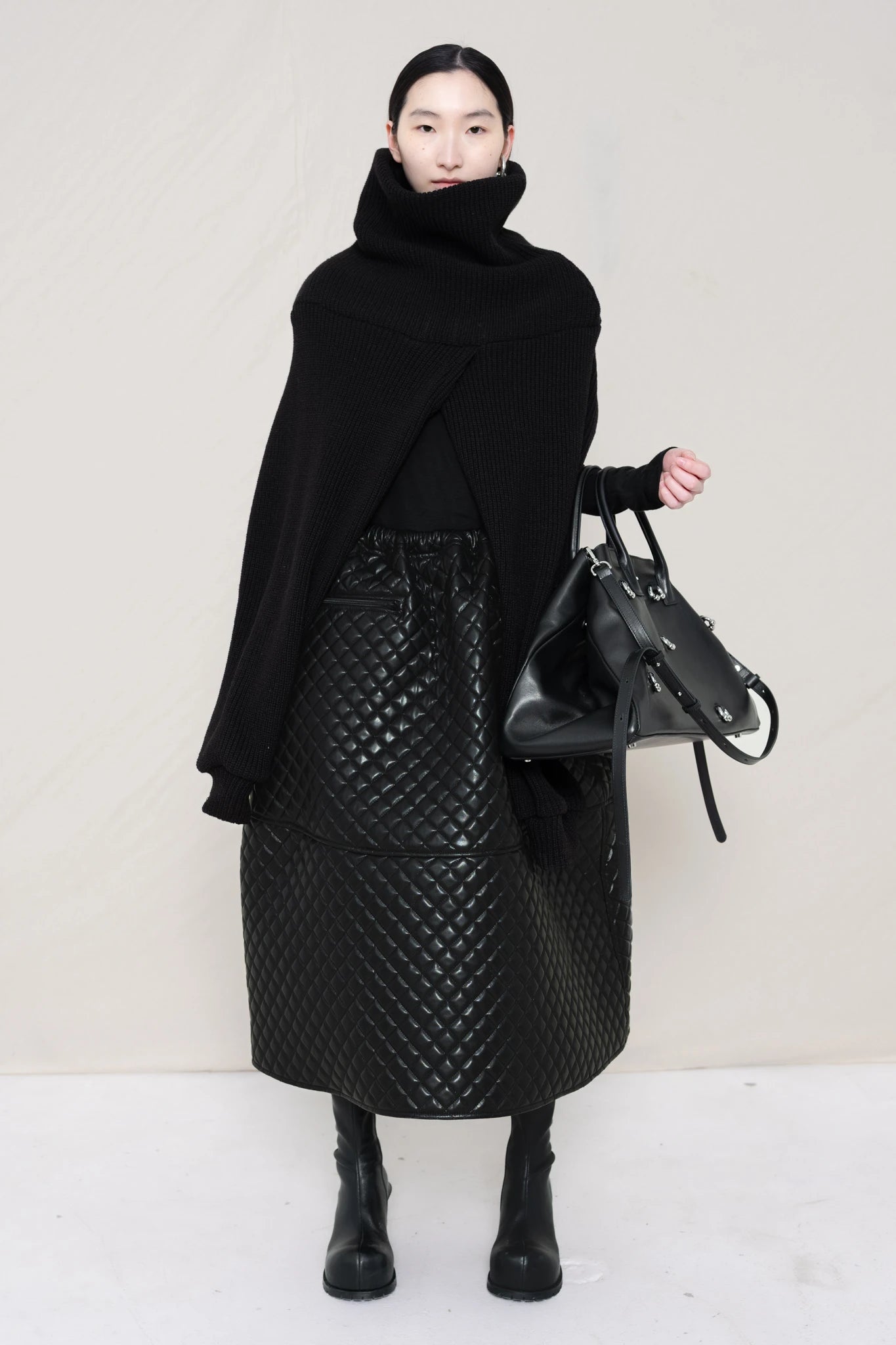 PERSON-WEARING-BLACK-CHUNKY-KNIT-TURTLENECK-THROW-OVER-WITH-BLACK-QUILTED-A-LINE-SKIRT-AND-BLACK-COWHIDE-DOUBLE-HEELED-BOOTS-HOLDING-BLACK-A-LINE-SHOPPER-WITH-PIERCING-DETAIL-FRONT-VIEW