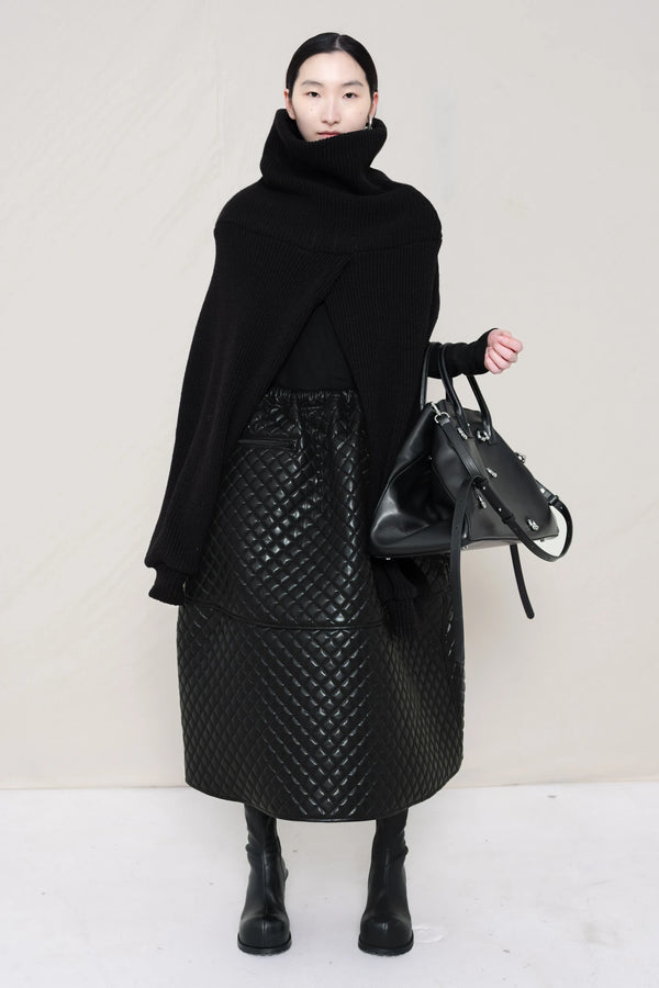 PERSON-WEARING-BLACK-CHUNKY-KNIT-TURTLENECK-THROW-OVER-WITH-BLACK-QUILTED-A-LINE-SKIRT-AND-BLACK-COWHIDE-DOUBLE-HEELED-BOOTS-HOLDING-BLACK-A-LINE-SHOPPER-WITH-PIERCING-DETAIL-FRONT-VIEW